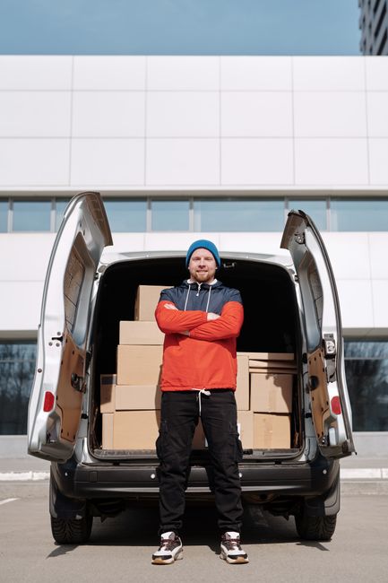 A man with a beard, wearing a blue beanie hat, red and navy jacket, black trousers, and sneakers, stands with arms crossed in front of an open van loaded with multiple stacked cardboard boxes. The van is parked outside a modern white building with large glass windows. The loading area is paved, and natural daylight illuminates the scene. The boxes are wrapped in plastic or tape, indicating they are packed for a home relocation or furniture transport. This image depicts the loading process of items during a professional house removal, with the man symbolising the staff from Man with Van Hounslow, providing efficient packing and moving services.