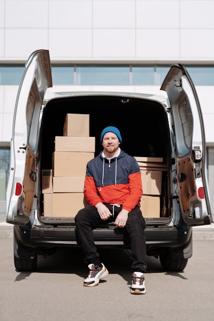 A man with a beard and a serious expression, wearing a red cap backwards, a two-tone beige and brown jacket over a checked shirt, and striped gloves, stands inside a moving van near the open rear door. Inside the vehicle, several cardboard boxes of varying sizes are stacked, some wrapped with packing tape. The van's interior features black upholstery and window curtains. The scene captures the loading process in a home relocation, with the man preparing to transport household items as part of a professional removals service. Natural lighting highlights the details of the packing materials and the man's attire, emphasizing the logistics involved in furniture transport and packing during house moves, as managed by companies like Man with Van Hounslow.
