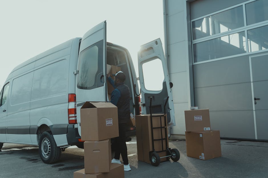 A man wearing a dark vest and hat is loading cardboard moving boxes into the back of a white van, parked outside a modern industrial building with large glass windows and a metal exterior. The scene captures the home relocation process, with several packed boxes arranged on the pavement near the van. A hand truck or dolly with a black handle is positioned nearby, ready to assist with furniture transport or cargo loading. The rear doors of the van are open, revealing additional packed items inside, while the sunlight casts a glow on the scene, emphasizing the professional removal operation. This image exemplifies the packing and loading stages involved in house removals, aligned with services provided by Man with Van Hounslow, as part of effective furniture transport and moving logistics solutions.
