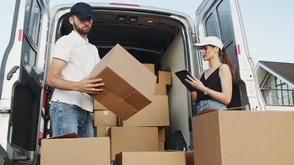 A man and a woman are standing at the rear of a white moving van, which has its doors open in a residential area with suburban houses visible in the background. The man, wearing a navy cap, white T-shirt, and jeans, is lifting and placing a medium-sized cardboard box into the van, part of a home relocation or furniture transport process. The woman, dressed in a white cap, black sleeveless top, and denim shorts, is holding a tablet and appears to be supervising or coordinating the packing and moving activities. Inside the van, several cardboard boxes of various sizes are stacked, some sealed with packing tape, alongside large sheets of protective foam or padded materials to safeguard fragile items during transit. The scene is lit with natural daylight, and the presence of packing materials, boxes, and lifting actions collectively illustrate a professional removal or house moving operation conducted by Man with Van Hounslow, focusing on efficient loading and transportation of household goods in the relocation process.