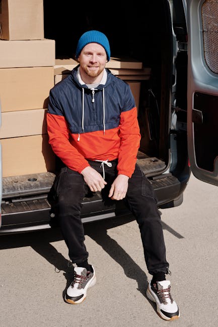 A man wearing a dark vest and hat is loading cardboard moving boxes into the back of a white van, parked outside a modern industrial building with large glass windows and a metal exterior. The scene captures the home relocation process, with several packed boxes arranged on the pavement near the van. A hand truck or dolly with a black handle is positioned nearby, ready to assist with furniture transport or cargo loading. The rear doors of the van are open, revealing additional packed items inside, while the sunlight casts a glow on the scene, emphasizing the professional removal operation. This image exemplifies the packing and loading stages involved in house removals, aligned with services provided by Man with Van Hounslow, as part of effective furniture transport and moving logistics solutions.