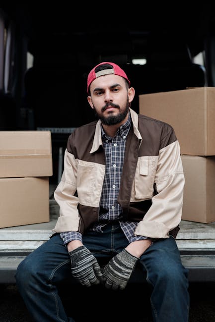 A young man with a beard, wearing a red baseball cap backwards, a brown and beige jacket over a checked shirt, and grey striped gloves, is sitting on the edge of a vehicle's loading area with his elbows resting on his knees. Behind him, the open rear of a van reveals several neatly stacked cardboard moving boxes. The boxes are standard-sized, made of brown corrugated cardboard, and some are sealed with tape, indicating they are packed for a home relocation. The interior of the van appears dark, with a glimpse of the vehicle's interior inside. The scene captures a moment during furniture transport and packing process, likely part of a moving service such as those offered by Man with Van Hounslow, supporting careful loading and logistical arrangements for house removals. The environment suggests a residential setting, with the van positioned on a pavement or driveway, ready for transportation of household belongings.