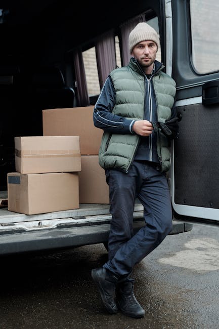 A man wearing a light grey knit beanie, a green padded vest over a navy blue hoodie, and dark trousers is standing next to the open rear doors of a black van used for house removals. Inside the van, there are several cardboard boxes of various sizes, some stacked on top of each other, and a large plain cardboard box positioned behind the man. The boxes are sealed with packing tape and appear ready for home relocation or furniture transport. The man is holding a black strap or restraint, possibly used to secure items during loading, with a neutral facial expression. The scene takes place outdoors on a paved driveway or street, with diffused natural lighting. This image illustrates the loading process involved in packing and moving services, supporting the services offered by Man with Van Hounslow for efficient removal logistics.