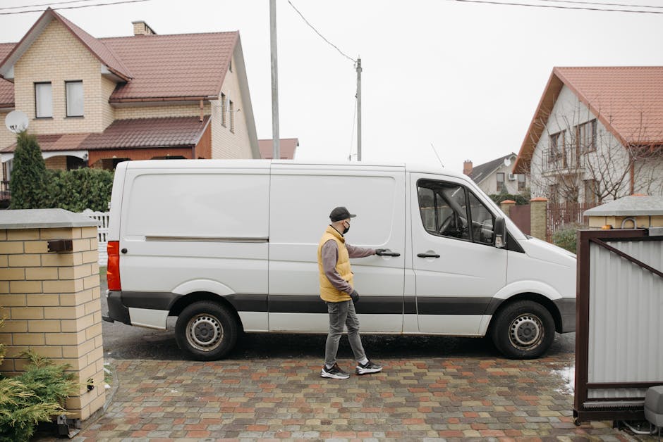 A man wearing a face mask, grey trousers, a grey long-sleeve top, and a yellow vest, stands outside a residential property, opening the sliding door of a white delivery van parked on a paved driveway. The van, used for house removals and furniture transport, has a large cargo area visible through the open sliding door. The driveway is bordered by brick walls and a small garden area with green shrubbery. In the background, there are two detached houses with pitched roofs covered in red tiles, some with visible windows, balconies, and chimneys. The scene is set during daylight in overcast weather. The image reflects a home relocation process, involving loading packing materials and furniture into the van, coordinated by a professional removals service such as Man with Van Hounslow, facilitating efficient packing and moving logistics for a household.