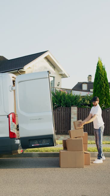 A man wearing a white t-shirt and jeans is standing on a paved residential street, loading cardboard boxes into the open rear doors of a white moving van. The van is parked close to a house with a sloping roof and a chimney, with a wooden fence and greenery in the background. The boxes, made of brown cardboard with visible packing tape, are arranged on the ground beside the man, who appears to be organizing them for a home relocation or furniture transport. Inside the van, partially visible through the open door, are more packed boxes and some wrapping materials. The scene is lit by natural daylight, emphasizing the orderly process of packing and moving goods in preparation for a house removal, with the presence of equipment such as the van and sturdy cardboard boxes indicating a professional moving service.