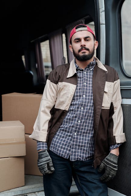A man wearing a face mask, grey trousers, a grey long-sleeve top, and a yellow vest, stands outside a residential property, opening the sliding door of a white delivery van parked on a paved driveway. The van, used for house removals and furniture transport, has a large cargo area visible through the open sliding door. The driveway is bordered by brick walls and a small garden area with green shrubbery. In the background, there are two detached houses with pitched roofs covered in red tiles, some with visible windows, balconies, and chimneys. The scene is set during daylight in overcast weather. The image reflects a home relocation process, involving loading packing materials and furniture into the van, coordinated by a professional removals service such as Man with Van Hounslow, facilitating efficient packing and moving logistics for a household.