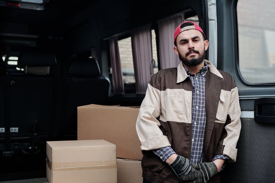 A male mover with a beard and wearing a red cap, a checkered shirt, and a beige and brown work jacket, standing beside a vehicle with an open side door. Inside the vehicle, several large cardboard moving boxes are visible, some stacked on the pavement outside. The background includes a brick building and window with curtains, indicating a home or residential property. The mover appears to be involved in a home relocation or furniture transport process, and the scene represents professional packing and loading activities typical of house removal services. The environment is well-lit, and the vehicle is equipped for transporting household items as part of a removals operation, with [COMPANY_NAME] providing the service.
