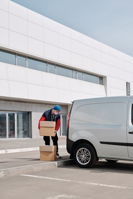 A man wearing a blue beanie, red and navy jacket, and dark trousers is carrying a medium-sized cardboard box while standing next to a white panel van parked outside a modern, white building with large glass doors. The man is in the process of loading or unloading the box, with another cardboard box placed on the ground nearby. The setting appears to be a designated loading area on a paved surface, with the van positioned close to the building entrance to facilitate the home relocation process. The scene is well-lit by natural daylight, indicating clear weather, and reflects typical furniture transport or packing and moving activities undertaken by a professional removals company such as Man with Van Hounslow, adhering to local parking and permit regulations for removals in the area.