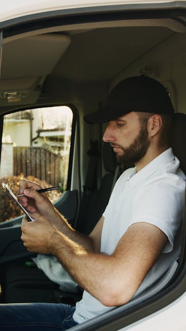 A male mover with a beard and wearing a red cap, a checkered shirt, and a beige and brown work jacket, standing beside a vehicle with an open side door. Inside the vehicle, several large cardboard moving boxes are visible, some stacked on the pavement outside. The background includes a brick building and window with curtains, indicating a home or residential property. The mover appears to be involved in a home relocation or furniture transport process, and the scene represents professional packing and loading activities typical of house removal services. The environment is well-lit, and the vehicle is equipped for transporting household items as part of a removals operation, with [COMPANY_NAME] providing the service.