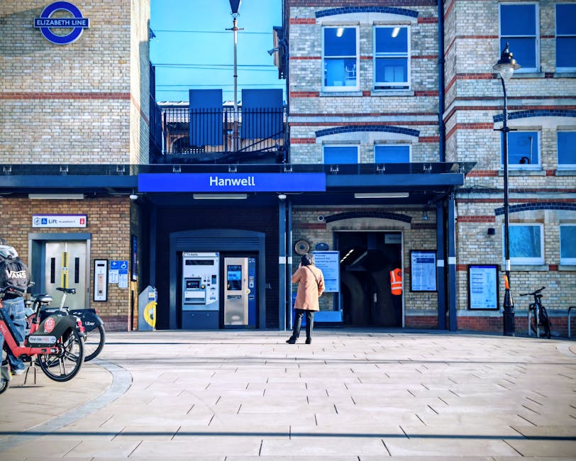 The image shows the exterior of Hounslow West underground station, part of the Elizabeth Line, featuring a brick building facade with multiple windows and a black awning displaying the station name. In front of the station, there is a paved pedestrian area with a woman wearing a beige coat, dark trousers, and black shoes, standing and looking towards the station entrance. To the left, a row of bikes including a VeloBike and a bike with a delivery box marked 'Uber' are parked on bike racks. The station entrance has a ticket machine, card payment terminals, and a lift access point, with an information board and signage indicating access routes. Overhead, a blue station sign displays “Hanwell”. A streetlamp is mounted on the corner of the building, and a small bicycle is secured to a black post on the right side of the scene. The lighting suggests daytime with clear visibility, and this setting relates to urban transport and access considerations for home relocation or moving services by companies such as Man with Van Hounslow.