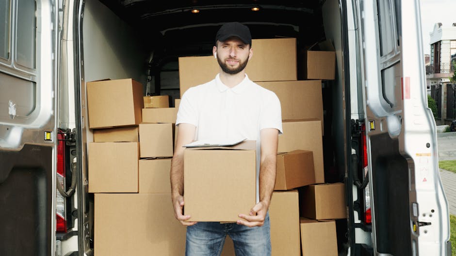 A man with a beard, wearing a white polo shirt, dark jeans, and a black cap, is standing inside the open rear of a white moving van, holding a cardboard box. The van is loaded with multiple cardboard boxes of various sizes, some stacked on top of each other and others placed on the floor, indicating a home relocation or furniture transport process. The interior of the van shows a clean, spacious cargo area with additional boxes in the background, ready for unloading or packing. The setting appears to be outdoors on a driveway or street, with a residential area visible in the background. The lighting is natural, illuminating both the man and the boxes, highlighting the careful handling involved in packing and moving activities. This scene is representative of professional removals services offered by Man with Van Hounslow, focusing on efficient packing and transport solutions for house moves near Hounslow West station.