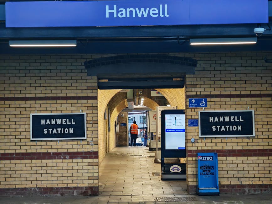 The image shows the exterior of Hounslow West underground station, part of the Elizabeth Line, featuring a brick building facade with multiple windows and a black awning displaying the station name. In front of the station, there is a paved pedestrian area with a woman wearing a beige coat, dark trousers, and black shoes, standing and looking towards the station entrance. To the left, a row of bikes including a VeloBike and a bike with a delivery box marked 'Uber' are parked on bike racks. The station entrance has a ticket machine, card payment terminals, and a lift access point, with an information board and signage indicating access routes. Overhead, a blue station sign displays “Hanwell”. A streetlamp is mounted on the corner of the building, and a small bicycle is secured to a black post on the right side of the scene. The lighting suggests daytime with clear visibility, and this setting relates to urban transport and access considerations for home relocation or moving services by companies such as Man with Van Hounslow.