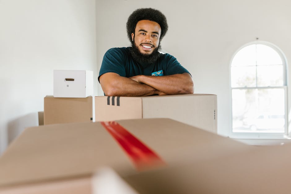 A smiling man with dark curly hair and a beard stands behind a collection of packed cardboard boxes inside a well-lit room with a large arched window in the background. The boxes are various sizes, some wrapped with protective plastic or packing tape, and are arranged on the floor, indicating they are prepared for a house removal or relocation. The man wears a dark blue t-shirt and has his arms crossed on top of the boxes, conveying confidence in the packing and moving process. The scene captures the preparation phase of furniture transport or home relocation, as part of the services offered by Man with Van Hounslow during packing and moving operations to Staines Road Hounslow.