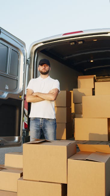 A man standing with arms crossed in front of the open rear doors of a white van parked on a pavement, surrounded by stacked cardboard boxes of various sizes, some sealed with packing tape and others open with flaps visible. Inside the van, additional boxes are visible, arranged in the cargo area. The scene is outdoors in natural daylight, related to home relocation or furniture transport, illustrating the loading process involved in house removals. The image reflects furniture packing, moving logistics, and transport preparations, aligning with the services offered by Man with Van Hounslow on their removals page.