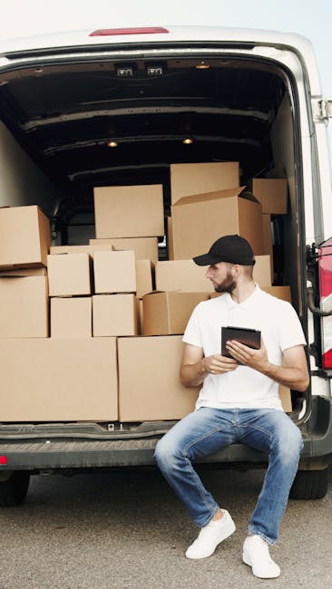 A young man wearing a white t-shirt, black cap, and blue jeans is sitting on the pavement beside an open moving van, which is filled with multiple cardboard boxes of various sizes, stacked neatly inside, some with flaps open or sealed with packing tape. The van appears to be parked outside a residential property, with the rear doors open, revealing the interior used for home relocation and furniture transport. The man is holding a tablet device, possibly reviewing packing lists or coordinating loading and unloading processes as part of a professional removals service. The scene is well-lit, with natural daylight, and various moving paraphernalia such as packing materials or straps may be present, indicating an active loading process managed by [COMPANY_NAME], specialized in house removals and moving logistics, supporting efficient packing and transport services in Hounslow.