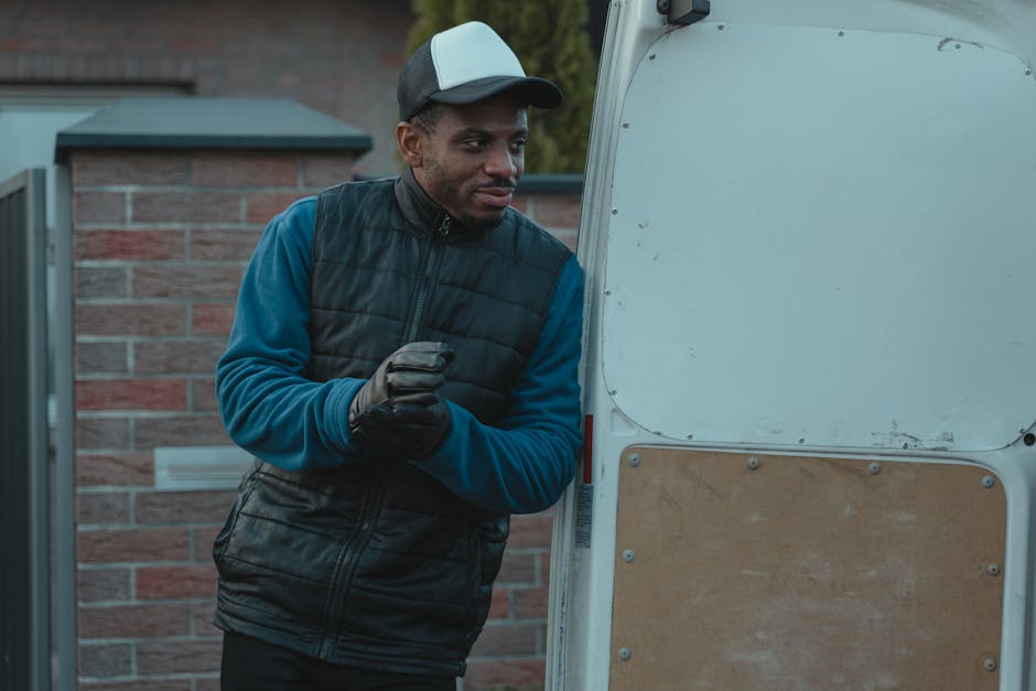 A man with dark skin wearing a black and grey baseball cap, a blue long-sleeve shirt, a black quilted vest, and black gloves is standing next to an open white van, which appears to be used for home relocation. The man is on the pavement outside a residential property with a brick wall and a gate visible in the background. Inside the van's open side door, cardboard boxes, padded furniture blankets, and pieces of packing material are visible, along with a wooden panel attached to the van's interior. The man looks focused and is holding a small item or perhaps adjusting equipment, suggesting he is in the process of packing or loading items for a furniture transport or moving service. The scene is set in daylight with natural lighting, and the environment indicates an ongoing packing and loading process typical of house removals. Man with Van Hounslow is providing a professional removal service, involving packing, loading, and transportation of household items.