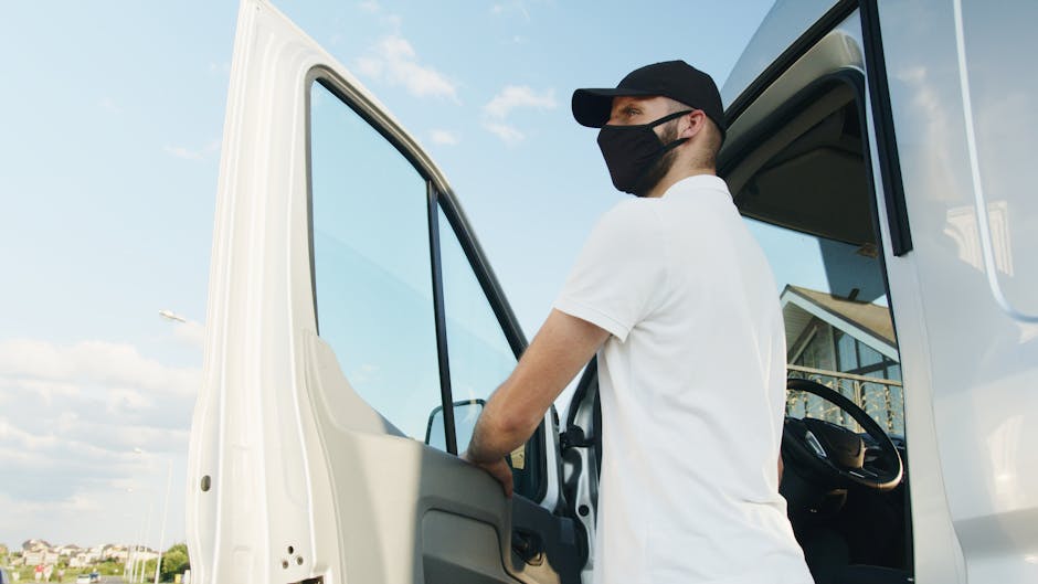 A young man wearing a white t-shirt, black cap, and blue jeans is sitting on the pavement beside an open moving van, which is filled with multiple cardboard boxes of various sizes, stacked neatly inside, some with flaps open or sealed with packing tape. The van appears to be parked outside a residential property, with the rear doors open, revealing the interior used for home relocation and furniture transport. The man is holding a tablet device, possibly reviewing packing lists or coordinating loading and unloading processes as part of a professional removals service. The scene is well-lit, with natural daylight, and various moving paraphernalia such as packing materials or straps may be present, indicating an active loading process managed by [COMPANY_NAME], specialized in house removals and moving logistics, supporting efficient packing and transport services in Hounslow.