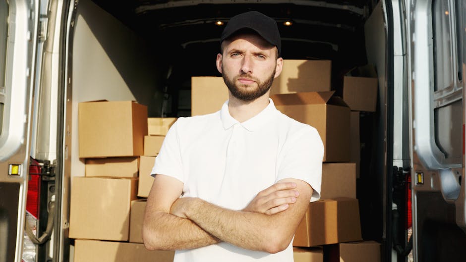 A young man with a beard, wearing a white polo shirt and a black cap, stands with his arms crossed inside the open rear of a moving van filled with cardboard boxes of various sizes, some wrapped in pl