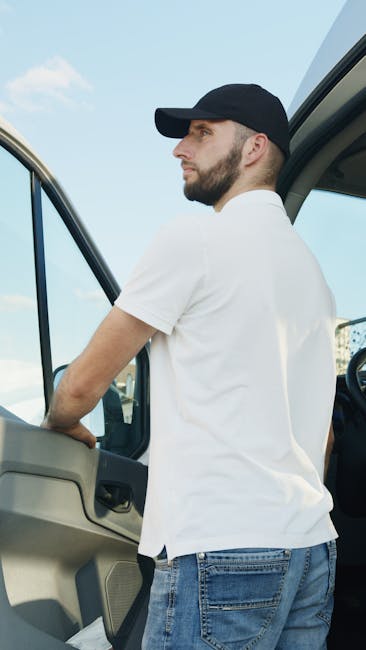A young man with a beard, wearing a white polo shirt and a black cap, stands with his arms crossed inside the open rear of a moving van filled with cardboard boxes of various sizes, some wrapped in pl